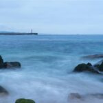Peaceful ocean view with rocks, waves, and a lighthouse in the distance.