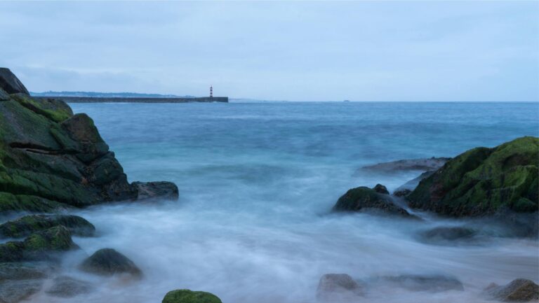 Peaceful ocean view with rocks, waves, and a lighthouse in the distance.