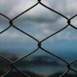 Close-up of a wire mesh fence with a blurred ocean and sky in the background, creating a pattern.