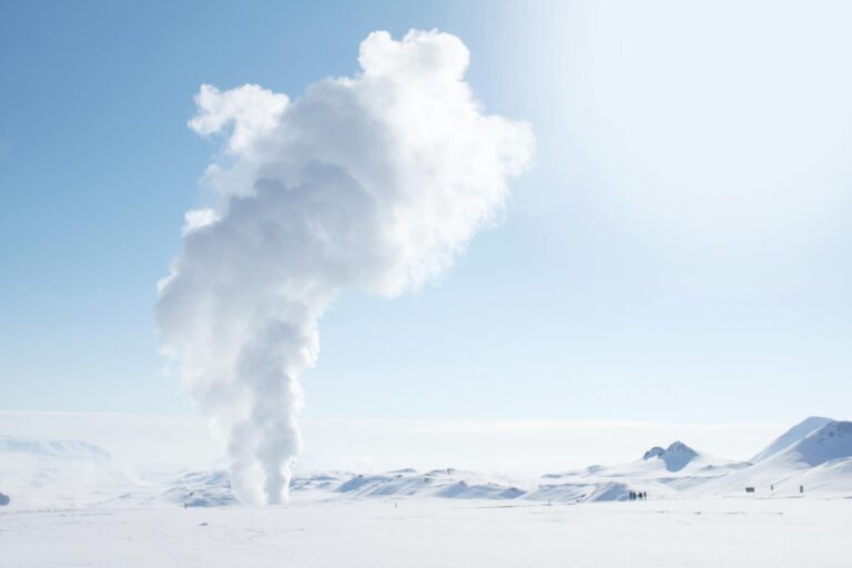 Snow-covered Icelandic landscape featuring geothermal steam plume against clear sky.
