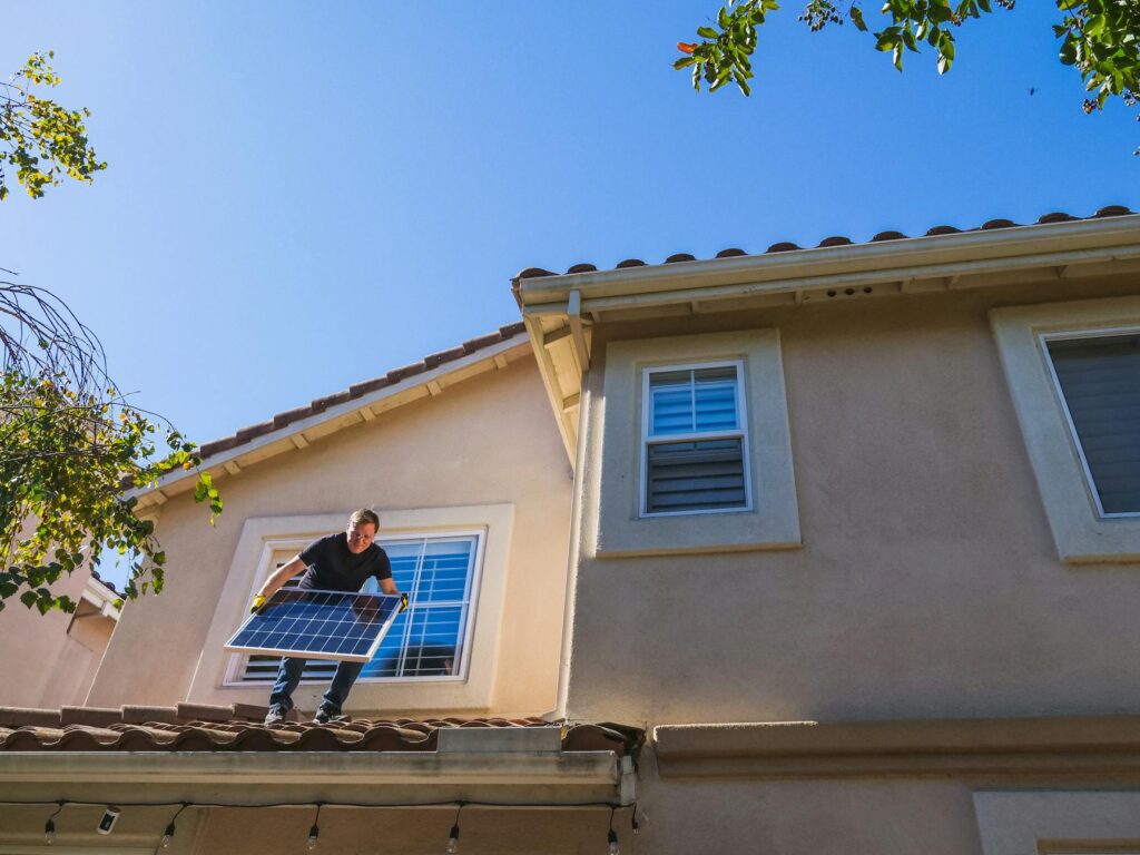 Man installing a solar panel on a rooftop under a clear blue sky, promoting renewable energy.