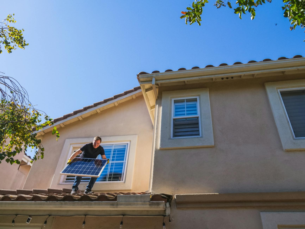 Man installing a solar panel on a rooftop under a clear blue sky, promoting renewable energy.