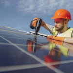 Low angle of serious bearded male electrician in hardhat and protective gloves installing solar photovoltaic panel system using drill