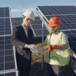 Two engineers inspecting solar panels at a renewable energy site under clear blue skies.