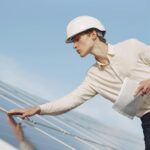A solar technician in a hard hat inspecting photovoltaic panels under clear skies.