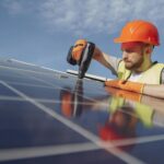 Low angle of serious bearded male electrician in hardhat and protective gloves installing solar photovoltaic panel system using drill
