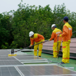 Three technicians in uniforms and hardhats clean rooftop solar panels in a renewable energy project.