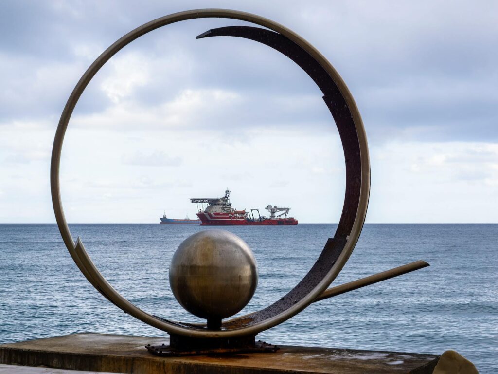 Artistic coastal sculpture frames a distant vessel on a serene ocean backdrop.