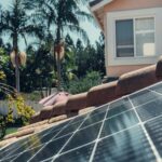 Close-up of solar panels on a rooftop with palm trees and a house in the background.