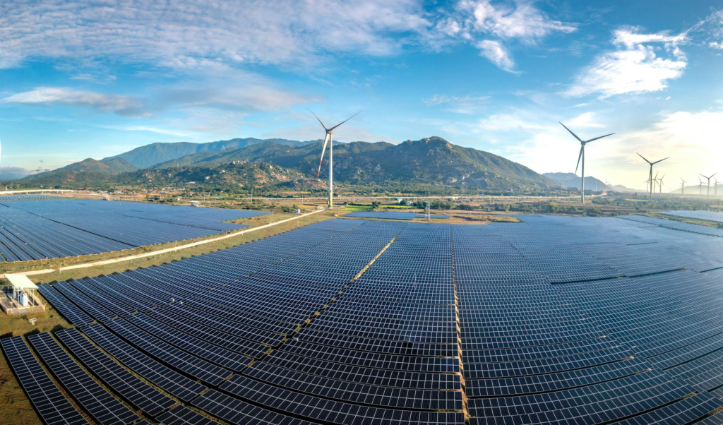 Aerial view of a vast solar farm with wind turbines against a mountainous backdrop on a sunny day.