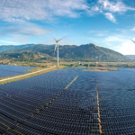 Aerial view of a vast solar farm with wind turbines against a mountainous backdrop on a sunny day.