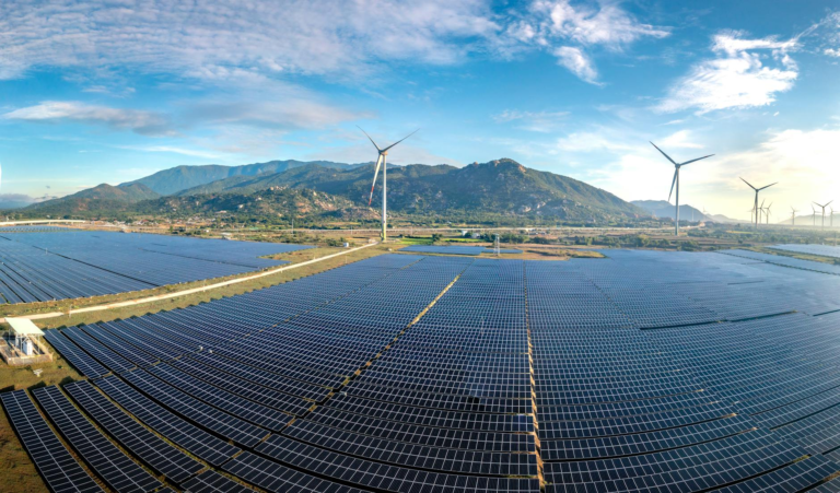 Aerial view of a vast solar farm with wind turbines against a mountainous backdrop on a sunny day.