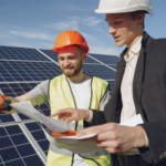 Low angle of young engineer and businessman in hardhats discussing project details against solar energy station