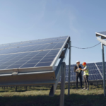 Supervisor in hardhat and formal wear discussing project with workman in uniform while standing with papers near modern solar panels under blue sky
