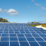 Expansive solar farm with rows of panels under clear blue sky and hills in the background.