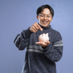 Smiling man holding a piggy bank and inserting a coin, representing savings and financial planning.