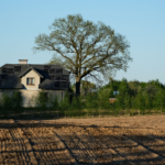 A rural house with solar panels surrounded by nature and farmland under clear blue skies.