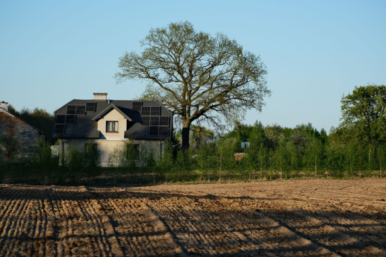 A rural house with solar panels surrounded by nature and farmland under clear blue skies.