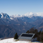 A stunning alpine view of snowy mountains and a solar-powered cabin under a clear blue sky.