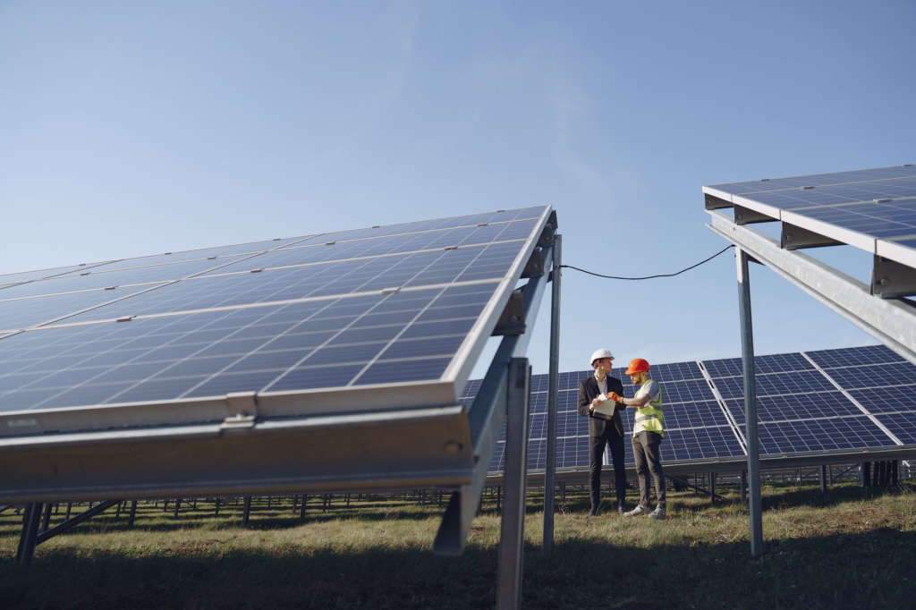 Supervisor in hardhat and formal wear discussing project with workman in uniform while standing with papers near modern solar panels under blue sky