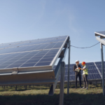 Supervisor in hardhat and formal wear discussing project with workman in uniform while standing with papers near modern solar panels under blue sky