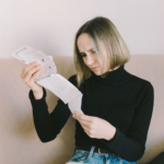 Concerned woman in black sweater examining bills on beige sofa indoors.