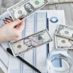 Close-up of a hand holding a dollar bill over financial documents and charts on a wooden table.