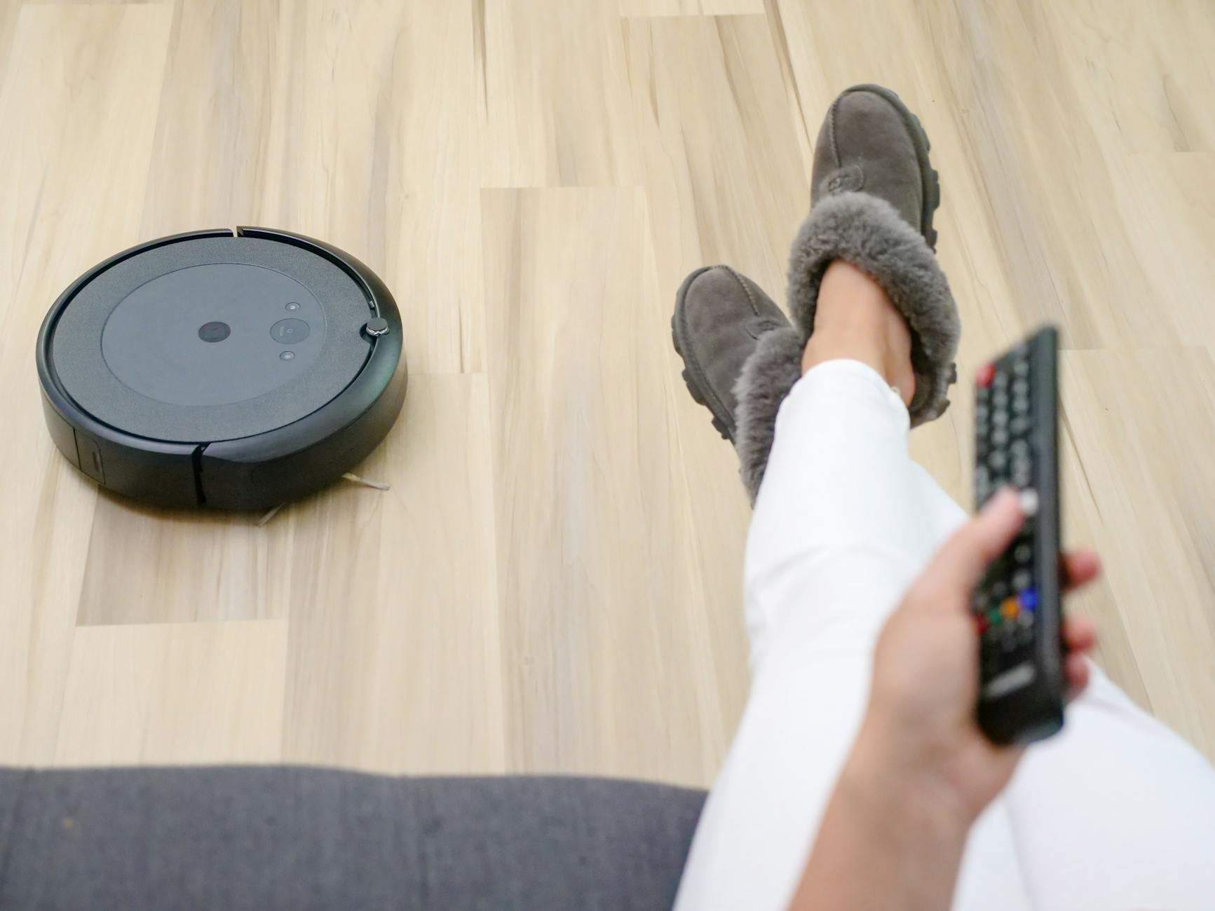 A person relaxing on a couch with a robotic vacuum cleaning the hardwood floor.